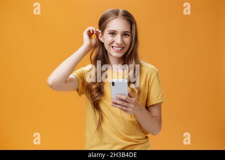 Waist-up-Aufnahme von niedlichen und freundlich aussehenden schönen jungen Mädchen mit welliger Frisur setzen Haarsträhne hinter dem Ohr halten Smartphone lächelnd breit mit Stockfoto