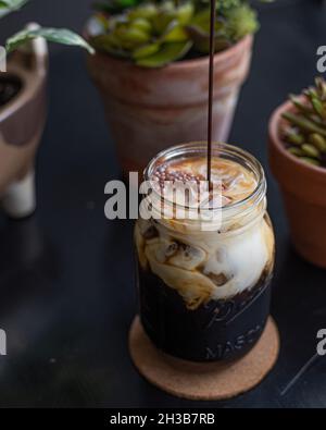 Kalter Brühkaffee mit Kokosmilch im Einmachglas, während Schokoladensirup aus dem Rahmen gegossen wird, umgeben von Sukkulenten und Pflanzen. Stockfoto