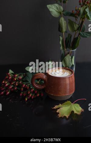 Ahorngewürzter Latte in handgefertigtem rostfarbenen Keramikbecher mit Tulpen-Latte-Kunst, umgeben von Herbstblättern auf schwarzer Kaffeetheke. Stockfoto