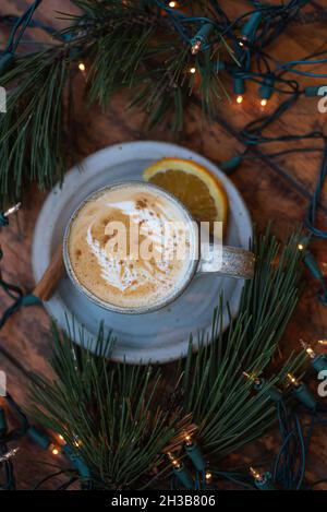 Urlaub gewürzte Zitrusfrüchte Latte mit Latte Kunst in handgefertigten grauen gesprenkelten Keramik Becher, umgeben von echten Kiefer Girlande und Weihnachtsbeleuchtung. Stockfoto