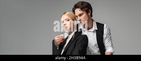 Stilvoller Mann, der die Zigarette einer blonden Frau aufhellt und die Kamera isoliert auf einem grauen Banner anschaut Stockfoto