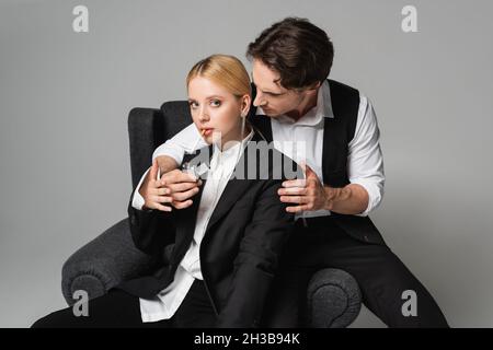 Eleganter Mann, der die Zigarette der blonden Frau aufhellt, die in einem Sessel auf Grau sitzt Stockfoto