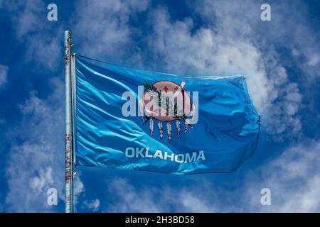 Flagge des Bundesstaates Oklahoma auf rostiger Stange, die im Wind vor einem wunderschönen, melierten, blau-bewölkten Himmel weht. Stockfoto