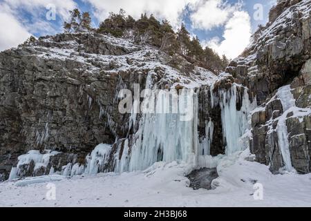 Ein großer Eisberg auf einem Berg aus zerklüfteten Felsen mit blauem Himmel und Wolken im Hintergrund. Der Berg ist mit langen Eiszapfen bedeckt, die hängen Stockfoto