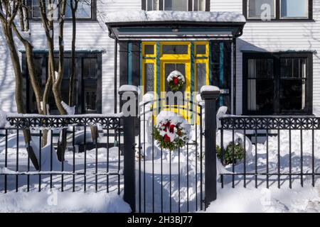 Der Eingang zu einem weißen Haus mit einer gelben Holz- und Glastür. Es gibt einen schmiedeeisernen Zaun aus schwarzem Metall und ein Tor im Garten. Ein Weihnachtskranz Stockfoto