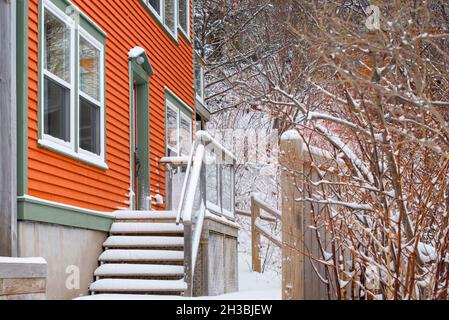 Die Außenfassade eines orangefarbenen Holzhauses mit grünen und weißen Zierleisten. Es gibt Stufen, die zum Eingang mit weißem Schnee führen. Stockfoto