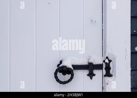 Eine weiße Holztür, die mit einem schwarzen schmiedeeisernen Verschluss geschlossen wurde. Der Mechanismus hat einen runden Griff. Oben liegt frischer weißer Schnee. Stockfoto