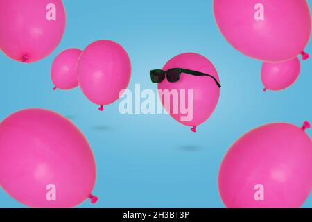 Pinker Ballon in Sonnenbrillen, der in der Luft schwimmt, mit anderen Ballons vor blauem Hintergrund. Minimales Konzept für Kreativität und Glück. Party-Stimmung, Stockfoto