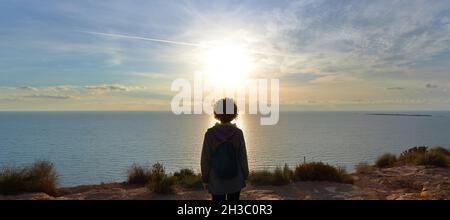 Panoramaaufnahme einer Frau von hinten, während die Sonne über dem Meer aufgeht Stockfoto