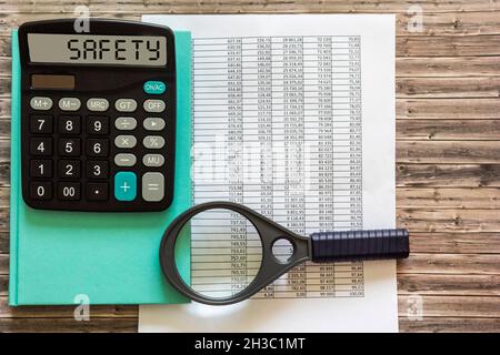 Sicherheitstext auf dem Taschenrechner-Display, grünem Notizblock, Lupe und Finanzberichten auf einem Holztisch. Stockfoto