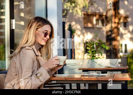 Schöne blonde Frau, die in einem Straßencafé Kaffee trinkt und an einem sonnigen Herbstnachmittag eine Sonnenbrille trägt. Stockfoto