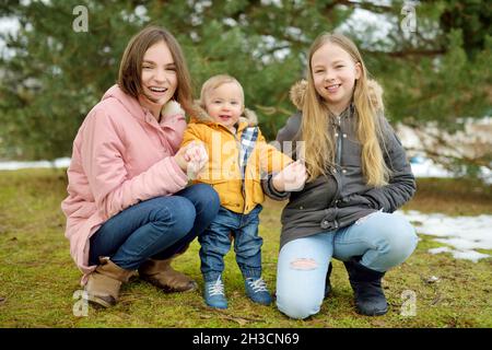 Zwei große Schwestern und ihr kleiner Bruder haben Spaß im Freien. Zwei junge Mädchen halten am Wintertag ihren Geschwisterjungen in der Hand. Kinder mit großer Alterslücke. Kind Stockfoto