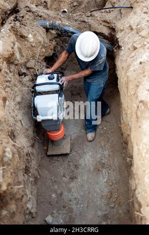 Bauarbeiter, der einen Bodenverdichter in einem tiefen Graben betreibt Stockfoto