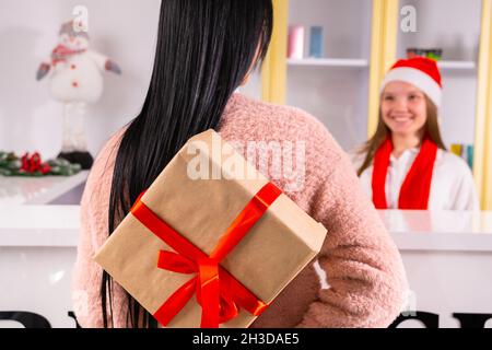 Rückansicht Foto einer jungen Brünette Frau, die die Geschenkbox mit dem roten Band hinter dem Rücken hält, um sie ihrem Kollegen in einem Weihnachtsmann-Hut und zu übergeben Stockfoto