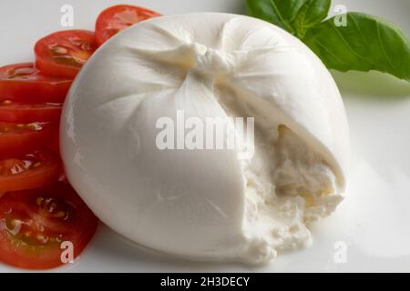 Frisches Stück Burrata-Käse Nahaufnahme auf weißem Hintergrund mit Tomatenscheiben und Basilikumblättern Stockfoto