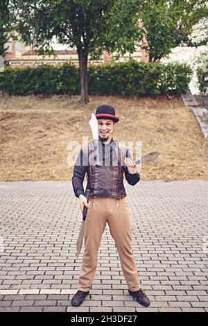 Full Body lustige männliche Darsteller in Kostüm lächelnd und jonglieren Messer, während auf dem Bürgersteig während der Show im Park stehen Stockfoto