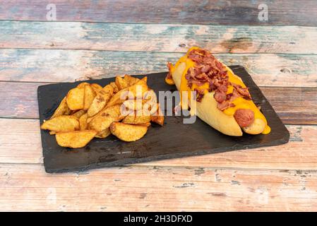 Hot Dog mit viel geschmolzenem Cheddar-Käse mit gebratenem Speck und hausgemachten Pommes auf schwarzem Schieferteller Stockfoto