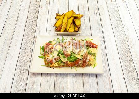 Argentinisches Schnitzel mit geriebenem Käse mit Parmesankäse-Flocken, serrano-Schinken und Rucola mit einer Garnierung aus in Keilen gerösteten Kartoffeln Stockfoto