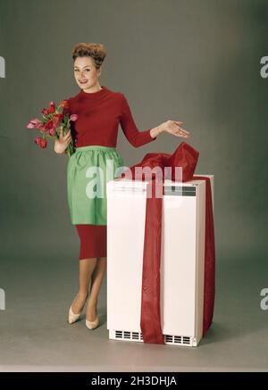 In den 1960er Jahren. Eine Frau in einem roten Kleid mit einer grünen Schürze steht neben einem Kühlschrank, in ein rotes Band gehüllt, als ob sie ihn als ersten Preis in einem Wettbewerb vorstellt. Schweden 60er Jahre Stockfoto