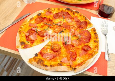 Köstliche italienische superdünne Kruste Pizza mit geräuchertem Speck und würzigen Pfefferoni in Scheiben geschnitten auf einem Teller an einem Restauranttisch Stockfoto