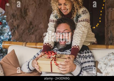 Weihnachtsfeiertage gibt Austausch Aktivität mit Mann und Frau zu Hause. Weibchen geben Geschenk bei erwachsenen Mann sitzen auf dem Sofa. Lady machen Sie eine Überraschung t Stockfoto