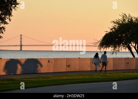 Das junge Paar hält sich romantisch die Hände und geht während des goldenen Sonnenuntergangs auf der Promenade mit der Mackinac Bridge, Lake Huron, Mackinac Island, Michigan, USA Stockfoto