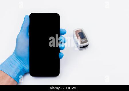 Die Hand eines Arztes in einem Latex-Gummihandschuh hält ein Mobiltelefon oder Smartphone mit einem Bildschirm vor dem Hintergrund des Pulsoximeters. Stockfoto