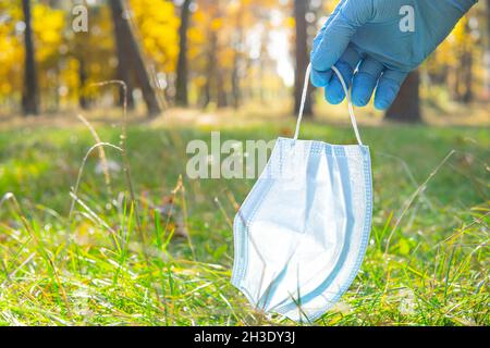 Nahaufnahme einer Hand, die einen Einweghandschuh trägt und eine weggeworfene einmal-Gesichtsmaske vom Boden im Stadtpark aufnimmt. Richtig sicher und umweltfreundlich Stockfoto