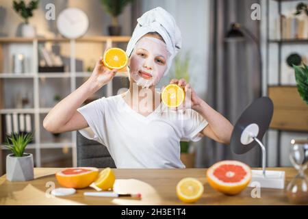 Hübsche weibliche Teenager mit kosmetischer Maske im Gesicht, die Orangenscheiben hält, während sie am Tisch sitzt. Hübsche junge Frau genießt Schönheitsroutine zu Hause. Stockfoto