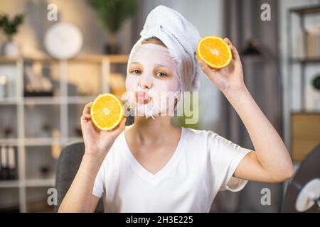 Positive junge Frau mit Badetuch auf dem Kopf posiert zu Hause mit Orangenscheiben in den Händen. Weibliche Teenager tun Vitaminmaske auf Gesicht für frische und gesunde Haut. Stockfoto