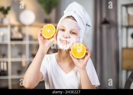 Charmante junge Frau, die Orangenscheiben in der Hand hält, während sie mit einer Maske im Gesicht und einem Badetuch auf dem Kopf vor der Kamera posiert. Konzept der Jugend- und Schönheitsbehandlung. Stockfoto