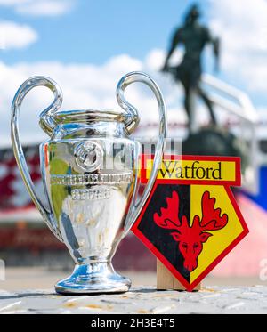 14. Juni 2021, Watford, Großbritannien. Watford FC Fußballverein-Emblem und der UEFA Champions League Cup vor der Kulisse eines modernen Stadions. Stockfoto