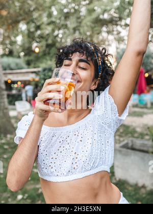 Positive ethnische Frauen trinken erfrischenden Cocktail und tanzen, während sie Party im Sommergarten genießen Stockfoto