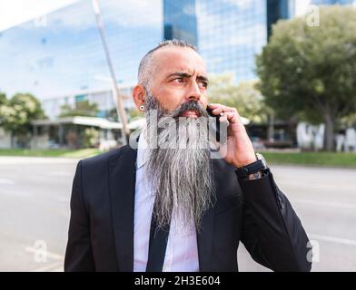 Selbstbewusster bärtiger Mann in elegantem Anzug, der Telefongespräche führt, während er in der Nähe der Straße mit modernen Gebäuden in der Stadt steht Stockfoto