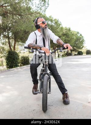 Selbstbewusster männlicher Hipster in weißem Hemd, der mit Kopfhörern Musik hört, während er auf dem Fahrrad auf der Straße mit grünen Bäumen sitzt Stockfoto