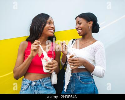 Glückliche afroamerikanische Freundinnen mit süßen Eisbechern, die sich gegenseitig angucken, während sie köstliche Desserts in der Nähe der Wand genießen Stockfoto