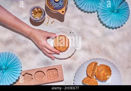Draufsicht auf Ernte anonyme Person mit traditionellen chinesischen Mondkuchen auf Teller auf Tisch mit Kräutertee und Backform platziert Stockfoto
