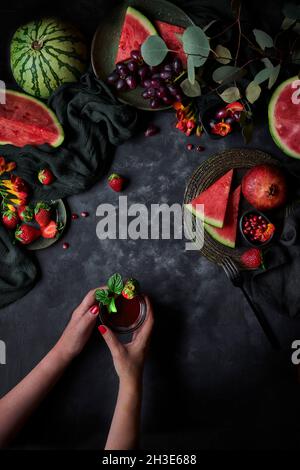 Draufsicht auf das anonyme Weibchen der Ernte mit einem Glas Getränk, das mit Erdbeer- und Basilikumblättern auf schwarzem Hintergrund mit Wassermelone und Granatapfel gekrönt ist Stockfoto