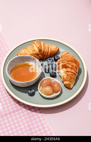 Köstliche Croissants auf Keramikplatte mit frischen Heidelbeeren und Marmelade auf rosafarbenem Tisch serviert Stockfoto