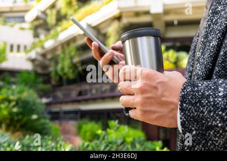 Seitenansicht des anonymen Mannes mit heißem Getränk in Tumbler-Textnachrichten auf dem Handy in der Stadt auf verschwommenem Hintergrund Stockfoto