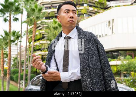 Gut gekleideter junger asiatischer Unternehmer in Krawatte schaut weg, während er auf der Straße gegen moderne Gebäude in der Stadt schlendert Stockfoto