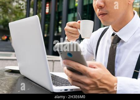 Beschnittene, nicht erkennbare ethnische männliche Unternehmerin mit Kaffee-Surfen im Internet auf dem Handy, während sie am städtischen Cafeteria-Tisch mit Laptop sitzt Stockfoto