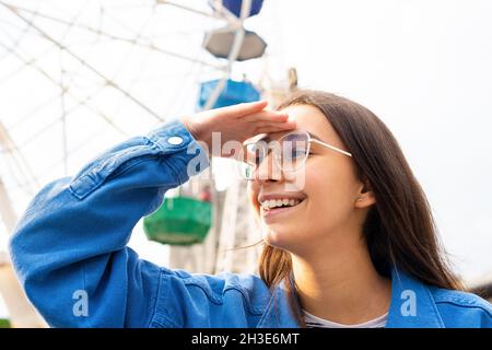 Junge lächelnde Brünette in Brille und Jeanshemd, die in die Ferne blickt und die Hand an der Stirn gegen das Riesenrad im Park hält Stockfoto