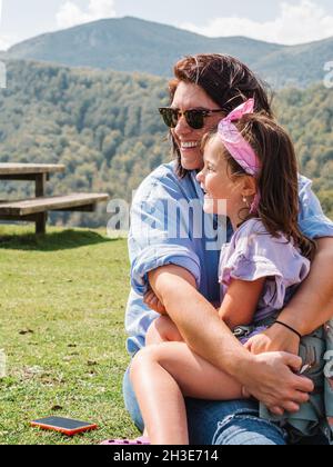 Positive Mutter in Sonnenbrillen mit niedlicher Tochter auf Runden, die auf grasbewachsenem Boden gegen Berg mit grünen Bäumen in der Natur sitzt Stockfoto