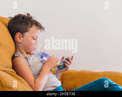 Inhalt kranker Junge, der Atemschutzmaske verwendet und Sauerstoff aus dem Inhalator atmet, während er zu Hause auf dem Sofa sitzt Stockfoto
