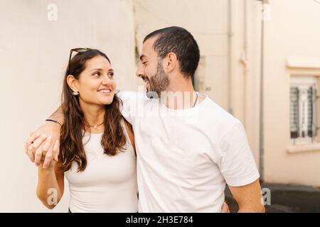 Fröhliches, verliebtes junges hispanisch-pärchen in lässiger Kleidung lachend, während sie die Straße entlang in die Stadt gehen Stockfoto