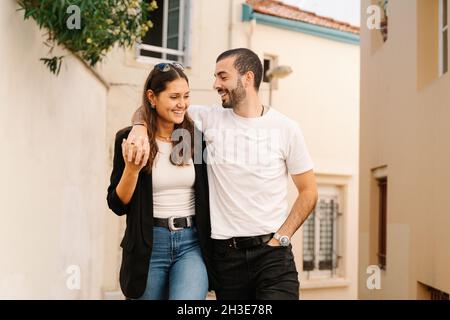 Aufgeregtes junges stilvolles hispanisch-panische Paar, das sich am sonnigen Tag auf einer schmalen Straße zwischen einem alten Wohnhaus kuschelt und lächelt Stockfoto