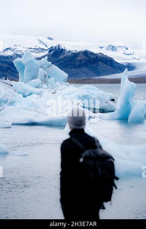 Rückansicht des anonymen Wanderers in warmer Oberbekleidung und Hut, der auf einem felsigen Hügel steht und die atemberaubende Landschaft des großen Gletschersees von Jokulsarlon duri bewundert Stockfoto