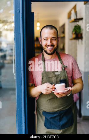 Positiver bärtiger Barista auf der Schürze mit einer Tasse heißen Kaffee in den Händen, der vor dem Eingang des modernen Cafés steht Stockfoto