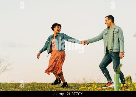 Die ganze Länge des positiven jungen multiethnischen Paares in legerer Kleidung hält Hände und lächelt, während er während eines romantischen Dates in CO auf einer Wiese spazierengeht Stockfoto